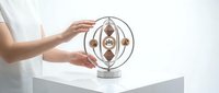 Woman hands protecting wooden kinetic sculpture with spinning spheres on white table demonstrating balance and motion in modern minimalist interior.