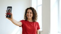 Young mixed-race woman in red shirt taking selfie with smartphone, smiling happily in bright modern interior with natural window light for social media.