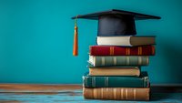Graduation cap with orange tassel placed on stack of vintage hardcover books against turquoise wall background on wooden surface. Academic achievement concept.