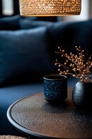 Moody interior with textured black ceramic cup and vase with dried flowers on dark table, illuminated by warm rattan pendant lamp against deep blue sofa background.