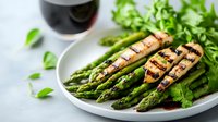 Grilled chicken breast slices served with fresh green asparagus spears on white plate, garnished with herbs and red wine glass in background.