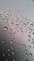 Water drops on glass surface creating natural pattern against blurred gray background, macro photography showing surface tension and condensation effect.