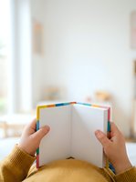 Child holding open colorful rainbow-edged notebook with blank pages in bright classroom setting, suitable for education, learning, creativity, and back-to-school projects.