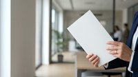 Businessman in dark suit holding blank white document or tablet in modern office corridor with colleagues in background and natural light from windows.