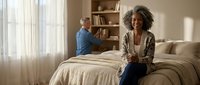 Mature African American woman with curly hair sitting on bed smiling while senior Caucasian man reads book by window in cozy bedroom interior.