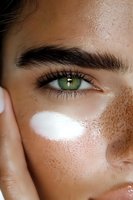 Close-up of bright green eye with natural freckles and moisturizer on young woman's face showing skincare and beauty routine details in macro photography.