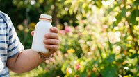 Farmer holding white bottle with supplement or organic product in garden with blooming flowers and green foliage background.