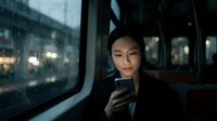 Young Asian woman using smartphone while sitting in vehicle at night, rain droplets on window, urban city lights visible in background, commuting or traveling by car or taxi.