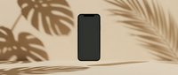 Smartphone with blank black screen on beige surface surrounded by tropical plant shadows and natural sunlight for app mockup and digital design projects.