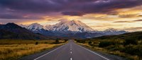Straight asphalt road stretching toward snow-capped mountain range under dramatic sunset sky with golden and purple clouds, suitable for travel and adventure themes.