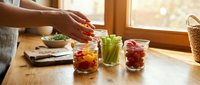 Woman preparing colorful fresh vegetable sticks in glass jars for healthy meal prep snacks on wooden kitchen counter near sunny window.