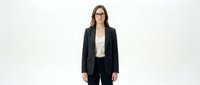 Young woman with long brown hair and glasses wearing a dark charcoal blazer over a cream-colored top with black trousers, standing against a white background for corporate and professional design use.