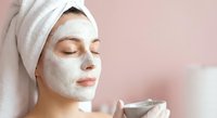 Young woman with white towel on head applying facial mask while holding cream jar for skincare routine and spa treatment at home.