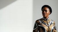 Young Black woman wearing ornate patterned shirt with gold and brown details, standing against white wall with natural light casting shadows, suitable for fashion, lifestyle, and portrait projects.