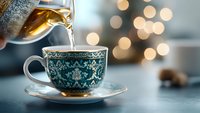 Steaming hot tea being poured into ornate vintage teacup with teal and white pattern against blurred bokeh lights background, creating cozy atmosphere.