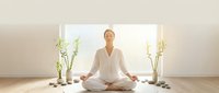 Young woman practicing meditation in lotus position wearing white clothing in bright minimalist room with bamboo plants and stones for wellness and mindfulness.