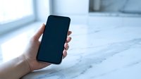 Hand holding black smartphone with blank screen on marble surface in bright modern office space for mockup and app presentation purposes.