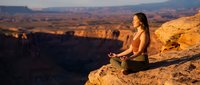 Young woman practicing yoga meditation on desert rock formation during golden hour sunset with canyon landscape background for wellness and mindfulness concepts.