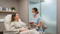 Female healthcare professional in scrubs consulting with relaxed patient in modern medical spa treatment room for wellness and beauty procedures.