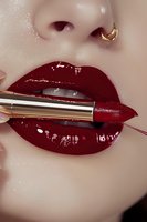 Close up of glossy dark red lips with gold nose piercing and metallic lipstick applicator, showing luxury cosmetics application on Asian female model.