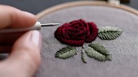 Detailed embroidery process of dark red rose with green leaves on gray fabric. Needlework artist creating floral design with needle and thread.