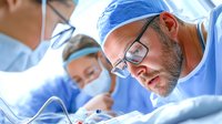 Caucasian male surgeon with glasses performing operation, focused on surgical procedure with medical team in operating room background. Medical care concept.
