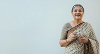 Senior woman with gray hair and South Asian ethnicity wearing traditional patterned saree, smiling warmly while holding fabric. Studio portrait against neutral background for lifestyle and cultural representation.