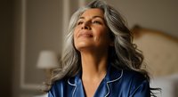 Mature woman with long gray hair and mixed ethnicity wearing navy blue medical scrubs with white piping, looking upward with contemplative expression in clinical healthcare setting.