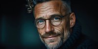 Mature Caucasian businessman with glasses and beard smiling warmly in professional portrait with dark background for corporate marketing materials.