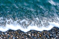 Aerial view of turquoise ocean waves crashing on pebble beach shore, creating white foam patterns against dark stones and crystal clear water. Perfect for travel themes.