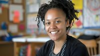 Young African American female teacher smiling warmly in colorful classroom setting with educational materials and books visible in background for learning concepts.