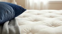 Cozy bedroom scene with navy blue pillow and cream quilted mattress, soft natural light from window, comfortable bedding arrangement for home decor and lifestyle content.