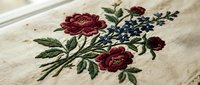 Traditional floral embroidery with burgundy roses and blue flowers on natural linen fabric, showcasing detailed needlework and textile craftsmanship.