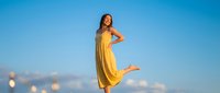 Young woman in flowing yellow dress dancing joyfully against clear blue sky background. Carefree summer lifestyle and freedom concept for wellness campaigns.