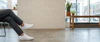 Modern minimalist office interior with person in dark pants and white sneakers sitting on chair, concrete floor, wooden bench, plant, large windows with city view.