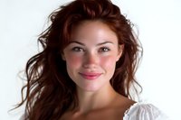 Young woman with flowing brown hair and natural makeup wearing white lace top smiles warmly at camera against light background, conveying joy and confidence.