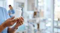 Healthcare professional holding blank white medication bottle in clinical setting with blurred medical staff background, suitable for pharmaceutical and wellness marketing.