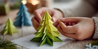 Hands making origami Christmas trees in green and blue paper, creating festive holiday decorations on wooden table with warm ambient lighting.