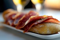 Spanish cured ham serrano slices served on fresh crusty bread rolls as traditional tapas appetizer with blurred wine glasses in background.