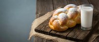 Fresh braided challah bread dusted with powdered sugar on rustic wooden cutting board with glass of milk, traditional Jewish bakery breakfast concept.