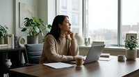 Young South Asian woman working at wooden desk with laptop, coffee cup, and notebook in bright modern office with large windows and potted plants, suitable for business, remote work, and professional lifestyle content.