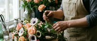 Florist arranging fresh cut roses and anemones in vase near window, hands holding peach blooms for floral design and decoration work.
