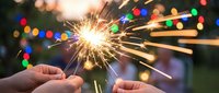 Hands holding lit sparkler with colorful bokeh lights in background, celebrating festive occasion or New Year's Eve party.