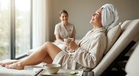 Relaxed woman in bathrobe and towel enjoying spa treatment while holding champagne glass with therapist in background at luxury wellness center.