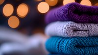 Cozy knitted sweaters stacked in purple, gray and turquoise colors with soft bokeh lights background creating warm autumn or winter mood for fashion design.