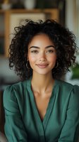 Young woman with curly dark hair and warm brown eyes wearing a green blazer, smiling at camera in modern office setting. Professional portrait for business, corporate, and lifestyle use.
