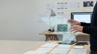 Businesswoman in dark blazer interacting with holographic financial data and charts floating above desk workspace with documents and laptop.
