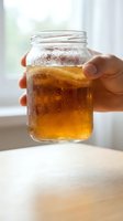 Hand holding glass jar filled with cold iced tea or cola beverage with condensation on exterior, refreshing drink on wooden table by bright window for beverage advertising.