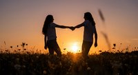 Two women holding hands in silhouette against golden sunset sky in wildflower field creating romantic friendship moment for wellness lifestyle content.
