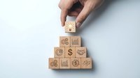 Hand placing wooden cube with lightbulb icon on pyramid of business symbol blocks representing innovation, growth strategy and entrepreneurial success.